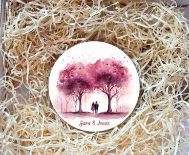 Runder Untersetzer mit romantischer Illustration von zwei Personen, die unter rosa blühenden Bäumen spazieren gehen. Unter dem Motiv stehen die Namen „Sara & Jonas“ in eleganter Schrift. Der Untersetzer liegt in einer Geschenkbox, umgeben von natürlichem